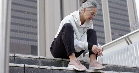 Mature woman tying running shoes on city steps wearing hoodie and fitness tracker