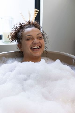 Laughing african american woman reclining in bubble bath with candlelight and reed diffuser
