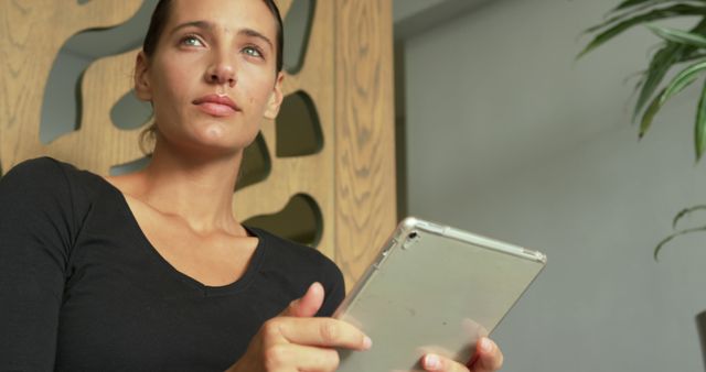 Young woman with brown hair using a digital tablet in modern relaxed indoor environment. The serene look on her face suggests deep concentration or thoughtful reading. Ideal for use in advertisements related to technology, modern lifestyle themes, or indoor settings focusing on peaceful and distraction-free environments.