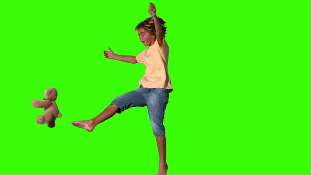 Boy in mid-air attempting to kick a teddy bear on green screen, perfect for digital compositing or advertising active play. Ideal for use in visual effects, online marketing, children’s product promotions, or playful themed content creation.