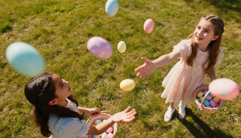 Two girls tossing pastel easter eggs in sunlit grass with wicker baskets, overhead view