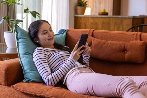 Asian woman relaxing on cozy couch using smartphone at home