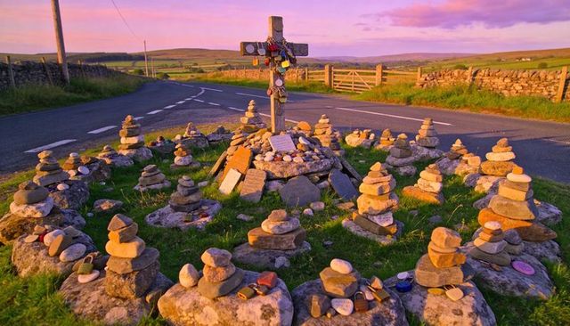 Wooden cross memorial at rural junction with padlocks, stacked stone cairns and painted pebbles