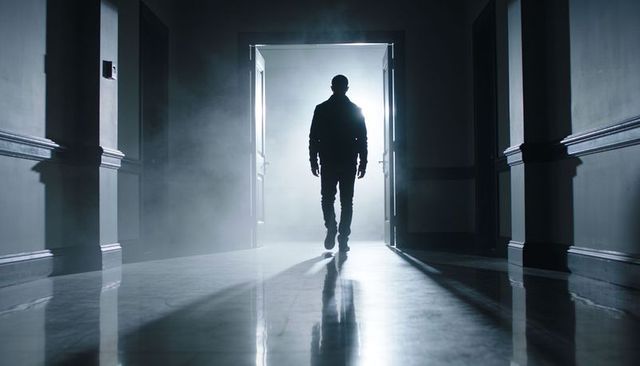 Silhouetted man walking through backlit double doors into foggy corridor, reflective floor