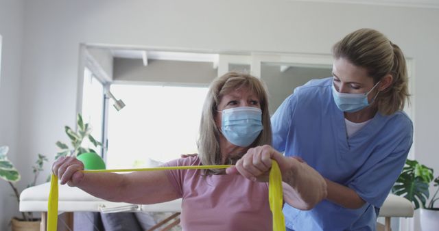 Senior woman doing rehabilitation exercises with assistance of a therapist at home. Both are wearing masks indicating a health-conscious environment. The therapist is guiding her with an exercise band. Ideal for illustrating senior care, at-home therapy sessions, physical rehabilitation, and safety protocols during pandemic situations.