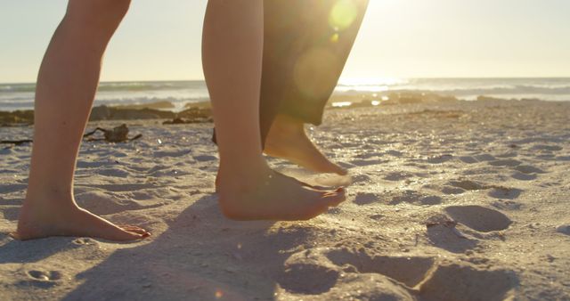 This image shows a close-up of two people walking barefoot along a sandy beach during sunset. The warm sunlight casts a soft glow on the sand, creating a tranquil and serene atmosphere. Suitable for use in travel advertisements, promotions for beach resorts, relaxation and wellness campaigns, and summer vacation content.