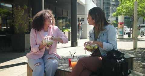 Two friends sharing fresh salads on urban bench, casual lunch and conversation