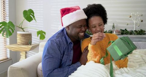 African American Couple Laughing While Opening Christmas Present on Cozy Living Room Couch