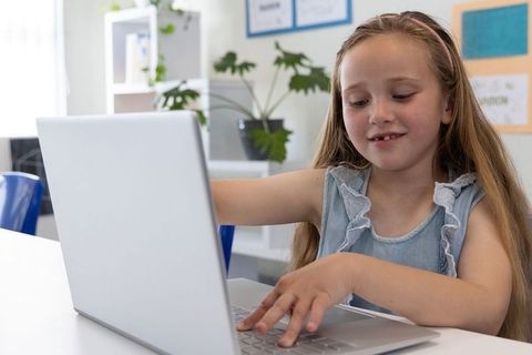 Elementary girl typing on laptop in bright classroom learning digital skills and studying