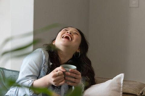 Joyful asian woman laughing while holding mug on sofa in cozy sunlit living room