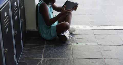 Schoolgirl sitting on corridor floor holding tablet, studying or reading in quiet hallway