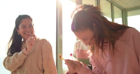 Young women testing lipstick and laughing in bright sunlit living room by tall windows