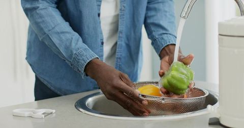 African american man washing bell peppers in colander at modern kitchen sink, rinsing produce