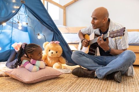 African american father playing guitar with daughter in cozy indoor teepee and lights
