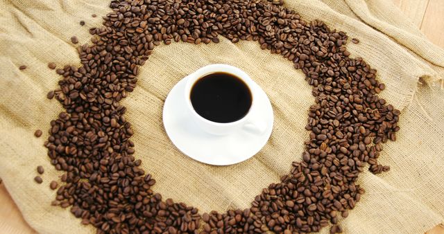 A cup of coffee sits surrounded by a circle of coffee beans on a burlap surface, with copy space. Its presentation suggests a focus on the rich culture and enjoyment associated with coffee drinking.