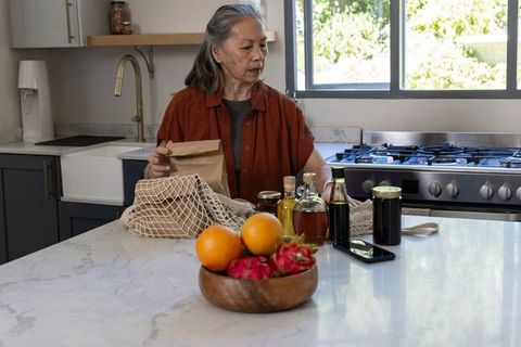 Senior asian woman unpacking groceries on marble kitchen island arranging jars and produce