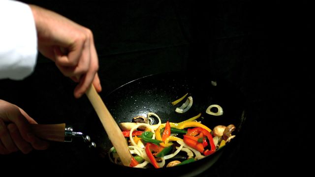 This image shows a chef expertly stirring a mix of colorful vegetables including red peppers, onions, and mushrooms in a wok, captured in slow motion. The dynamic motion and vibrant colors make this suitable for use in culinary blogs, cooking tutorials, or health-related content. It showcases the art of frying and can appeal to both professional chefs and cooking enthusiasts looking for vibrant culinary images.