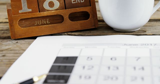 A wooden calendar set to June 10th is placed next to a white mug and a pen on top of a paper calendar, with copy space. It suggests a planning session or a reminder of an important date.