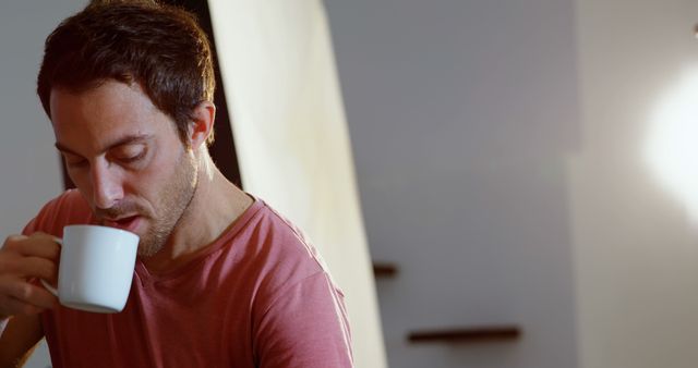 Young man enjoying coffee indoors during a quiet, relaxed moment. Perfect for use in materials related to relaxation, morning routines, contemporary lifestyle, leisure, and personal wellbeing.