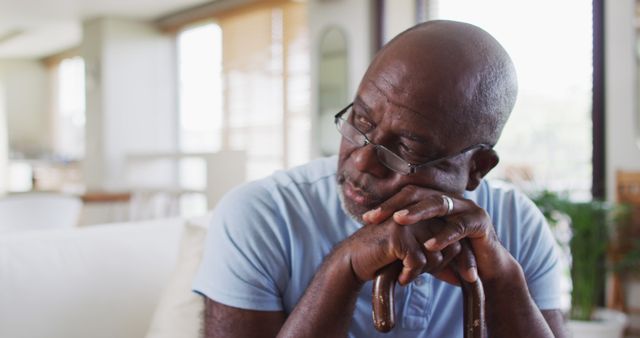 Elderly man is looking thoughtful, seated indoors. He is wearing glasses and lightly gripping a walking stick, giving an impression of contemplation and reflection. Ideal for use in healthcare, senior living, aging, mental wellness, and lifestyle-related projects.