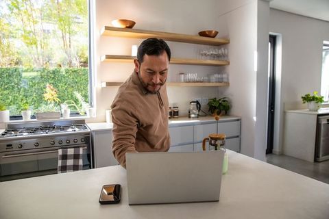 Asian man working on laptop at modern kitchen island with french press and greenery