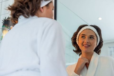 Woman smiling at mirror in white bathrobe practicing morning skincare ritual