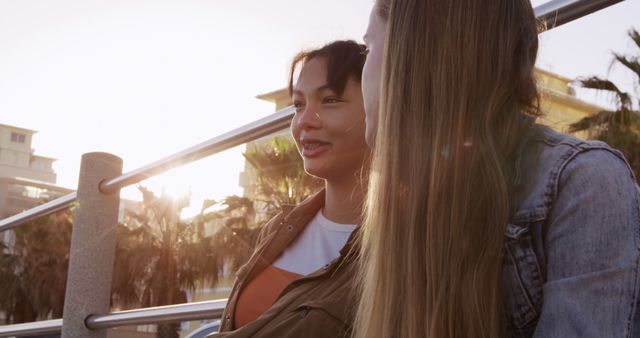 Two women chatting and enjoying each other's company outdoors during sunset, with palm trees in the background. Ideal for advertisements, social media promotions, blog posts about friendship, leisure, and outdoor activities.