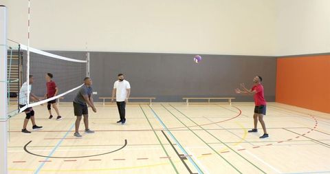 Diverse male volleyball team practicing indoor passing and receiving with coach observing on gym cou