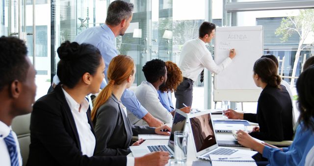 Diverse group of professionals are attentively listening to a colleague presenting information on whiteboard in a modern conference room. This could be useful for illustrating corporate meetings, teamwork, diversity in workplace, strategic planning sessions, and business presentations in promotional materials or reports.