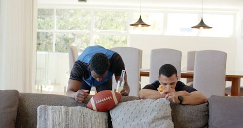 Diverse friends leaning on sofa reaching for football, eating pizza and holding beer