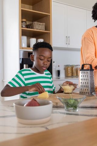 African american boy preparing shredded cheese with caregiver at modern kitchen island