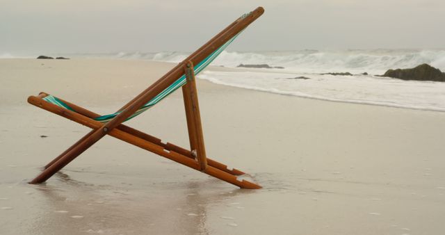 Empty wooden beach chair resting on quiet sandy shore by ocean's edge with gentle waves; provides serene setting. Great for concepts of solitude, relaxation, nature, vacations, and tranquility-oriented designs.