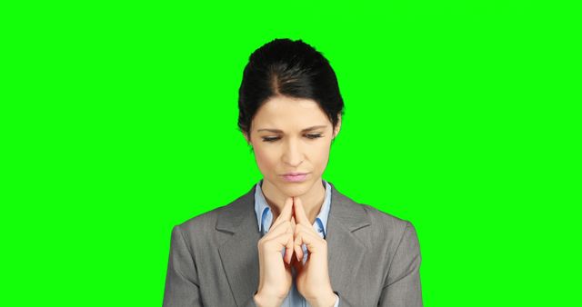 A middle-aged Caucasian businesswoman appears deep in thought, with her fingers pressed together, against a green screen background, with copy space. Her expression suggests she is contemplating a serious decision or problem.