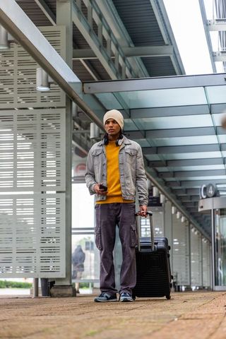 Young traveler waiting under glass canopy at transit terminal with suitcase and smartphone