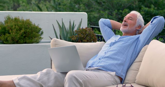 This image showcases an old man with white hair lounging comfortably on a patio chair, hands behind his head, and a smiling expression while his laptop rests on his lap. Taken against a backdrop of lush greenery and outdoor plants, this image can be used to illustrate concepts of remote work, relaxed lifestyle, enjoying retirement, and embracing nature. Perfect for advertisements, lifestyle websites, and articles focusing on senior living, digital nomad life, and healthy living.