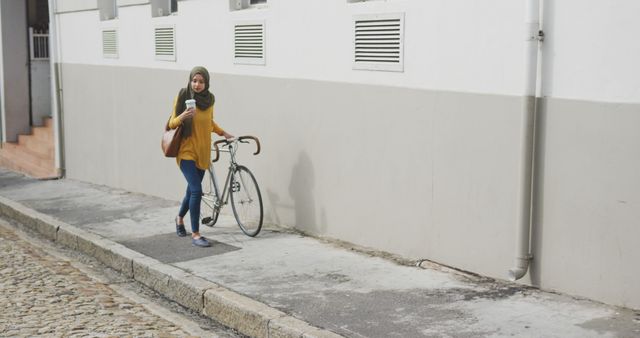 Biracial woman in hijab holding takeaway coffee and wheeling bicycle in quiet city street. City living, transport and modern urban lifestyle.