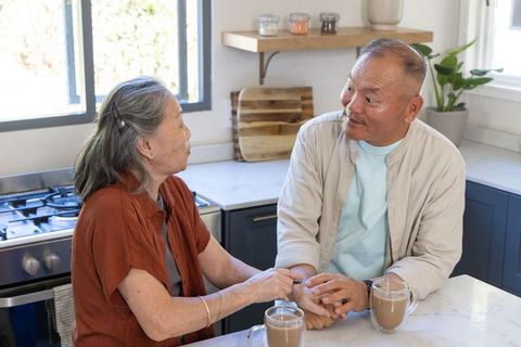 Senior asian couple holding hands and talking over coffee in bright home kitchen