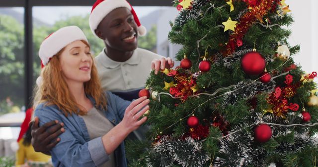Happy diverse couple wearing santa hats decorating christmas tree. Spending quality time at home together at christmas.