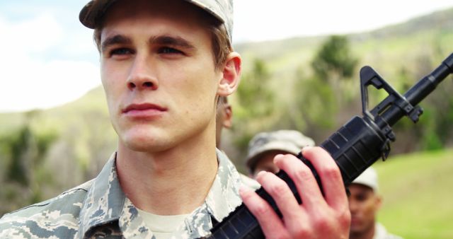 Young military soldier holding rifle outdoors in a field. Perfect for themes related to military training, armed forces recruitment, resilience, and duty. Can be used in articles, advertisements, and projects about military life, outdoor survival training, and youth in the armed forces.