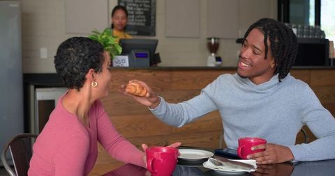 Young friends sharing croissant and coffee while smiling in cozy urban cafe