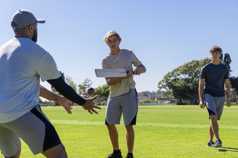 Men playing casual cricket in park, batter swinging flat bat while teammates catching