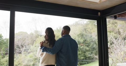 Couple embracing and gazing through floor-to-ceiling window at lush garden view