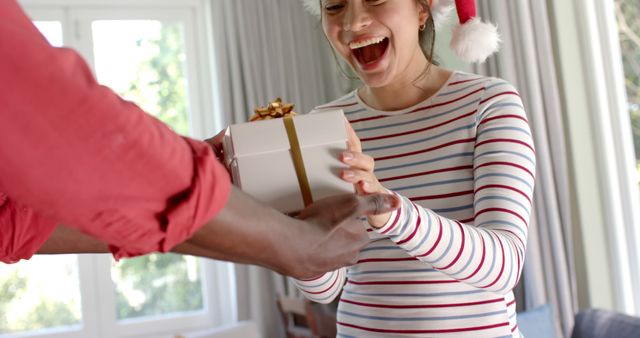 A woman wearing a striped shirt and Santa hat receiving a gift with excitement and joy. Perfect for holiday advertisements, Christmas card designs, festive marketing materials, or celebratory social media posts highlighting the joy of giving during the holiday season.