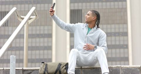 Young man taking selfie on urban ledge wearing gray hoodie holding smartphone duffel