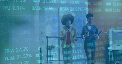 Two businesswomen walking and reviewing stock ticker overlay in modern office corridor