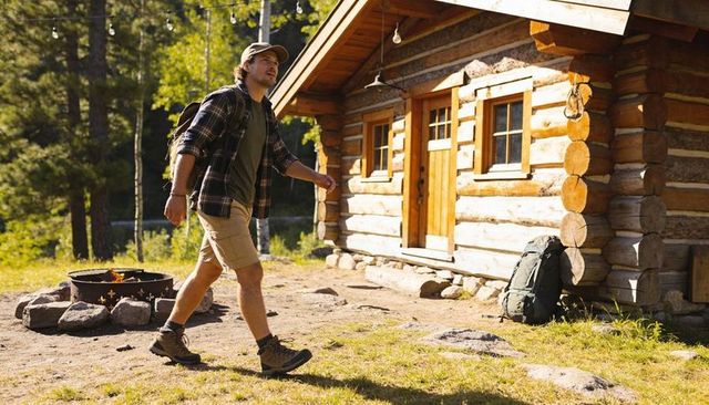 Solo hiker walking to rustic log cabin campsite with backpack and campfire in forest