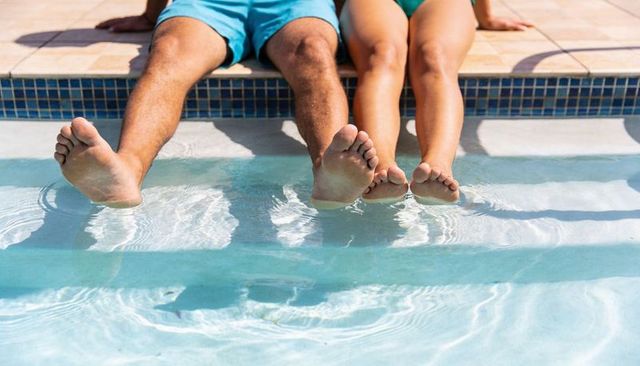 Couple sitting poolside dipping legs in sunlit water with mosaic coping and ripples