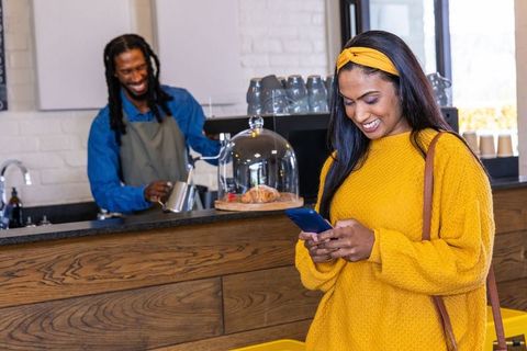Young african american woman checking smartphone at trendy coffee counter in mustard sweater