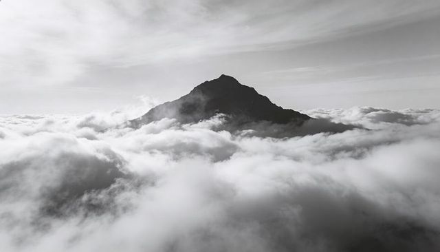 Rising jagged mountain peak piercing dense cloud sea monochrome aerial silhouette