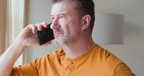 Middle-aged Man Talking on Smartphone by Window at Home